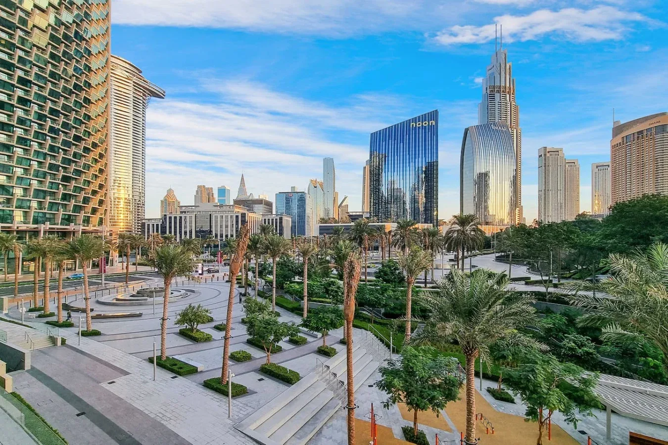 City skyline rises behind a landscaped plaza with palm trees and paved walkways.