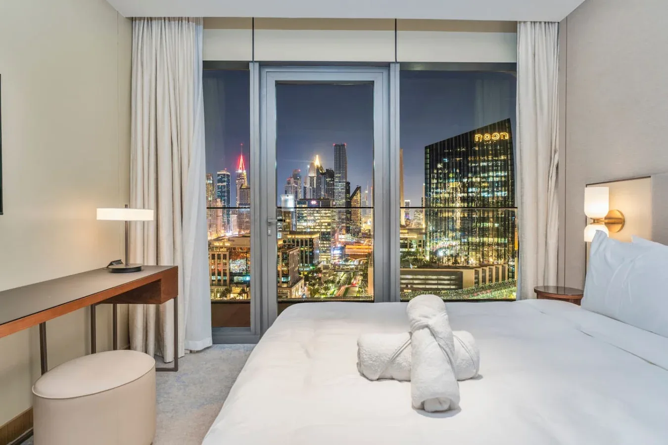 A bedroom with a large bed facing floor-to-ceiling windows overlooking a city at night.