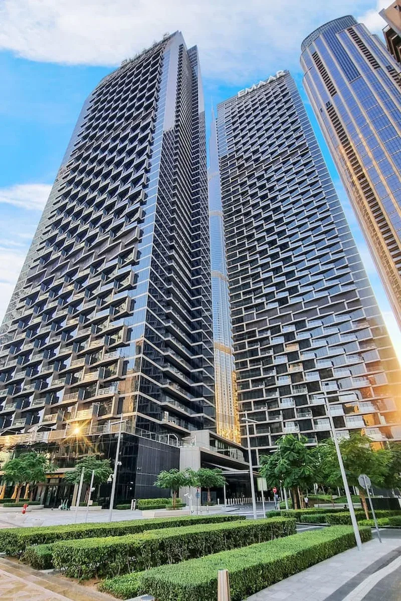 Tall modern buildings rise above landscaped greenery and a paved walkway.