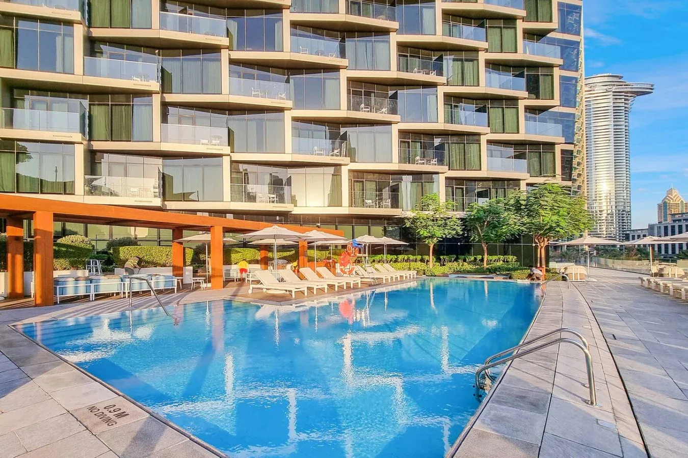 An outdoor swimming pool sits beside a modern building with balconies and lounge seating.