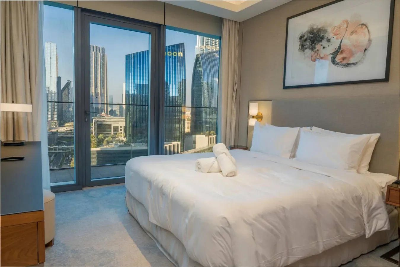 A bedroom with a large bed beside floor-to-ceiling windows overlooking city buildings.