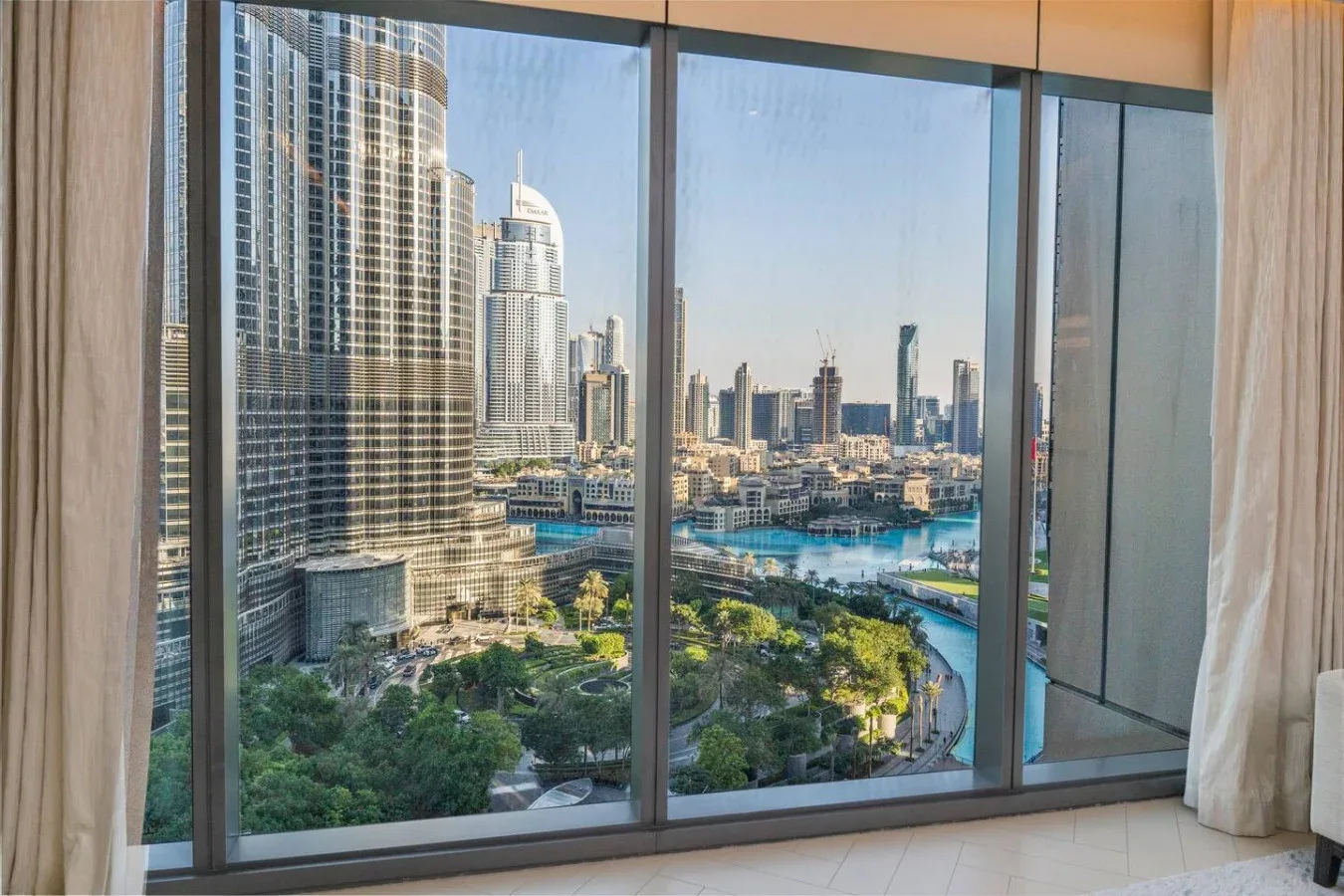 A high-rise room overlooks a city skyline, landscaped park, and turquoise water.