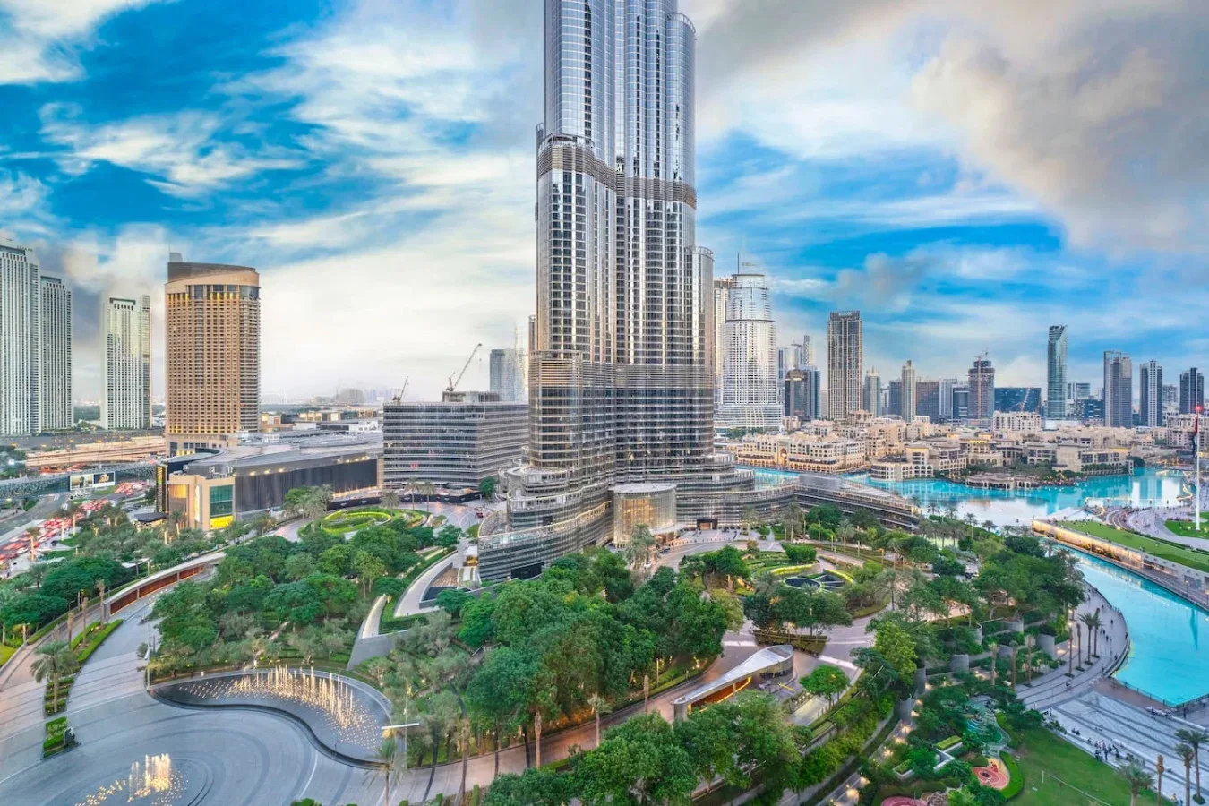 A city skyline surrounds a tall central tower, with landscaped greenery and a blue waterway in the foreground.