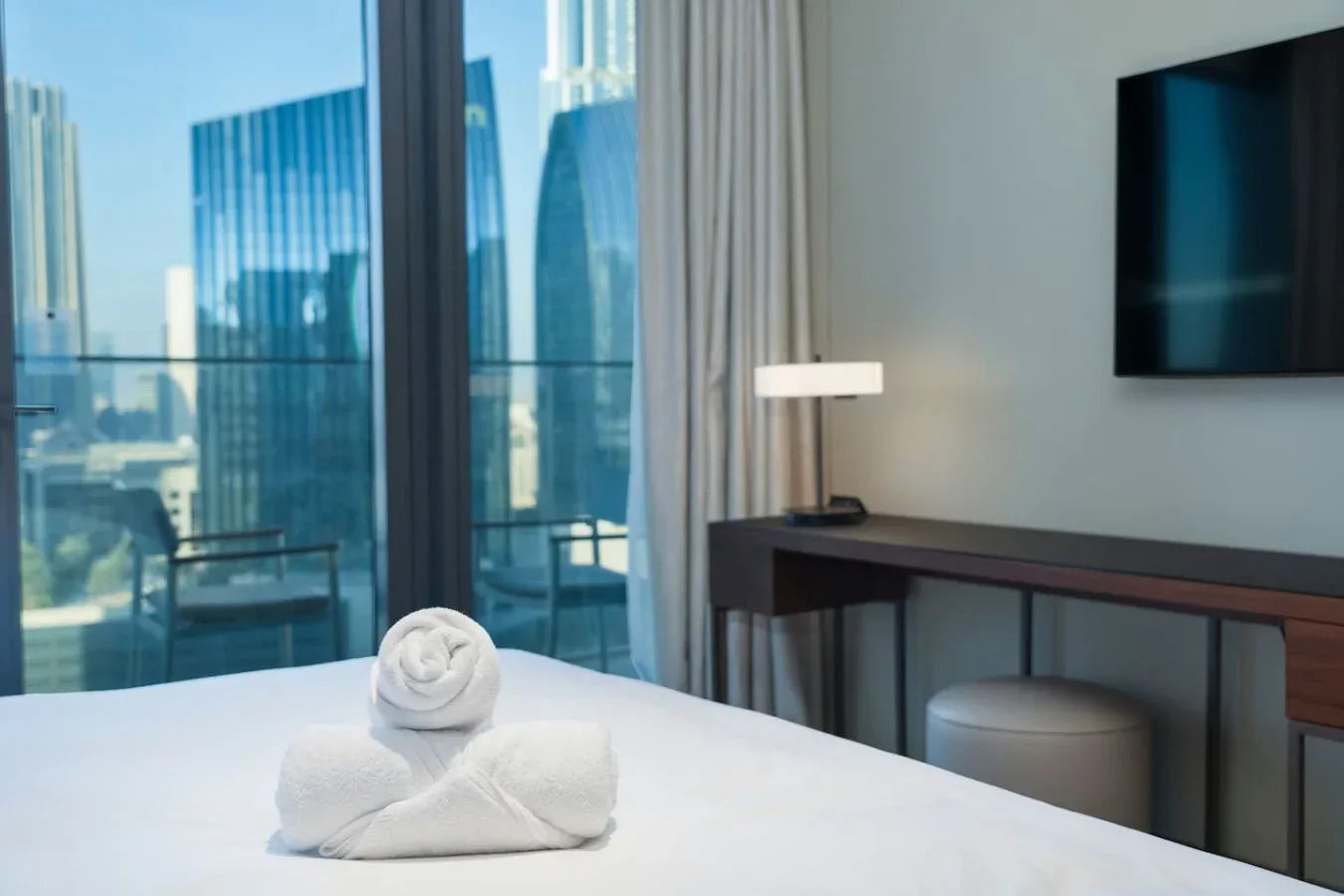 A bedroom with a neatly made bed, rolled towels, and a city view through the window.