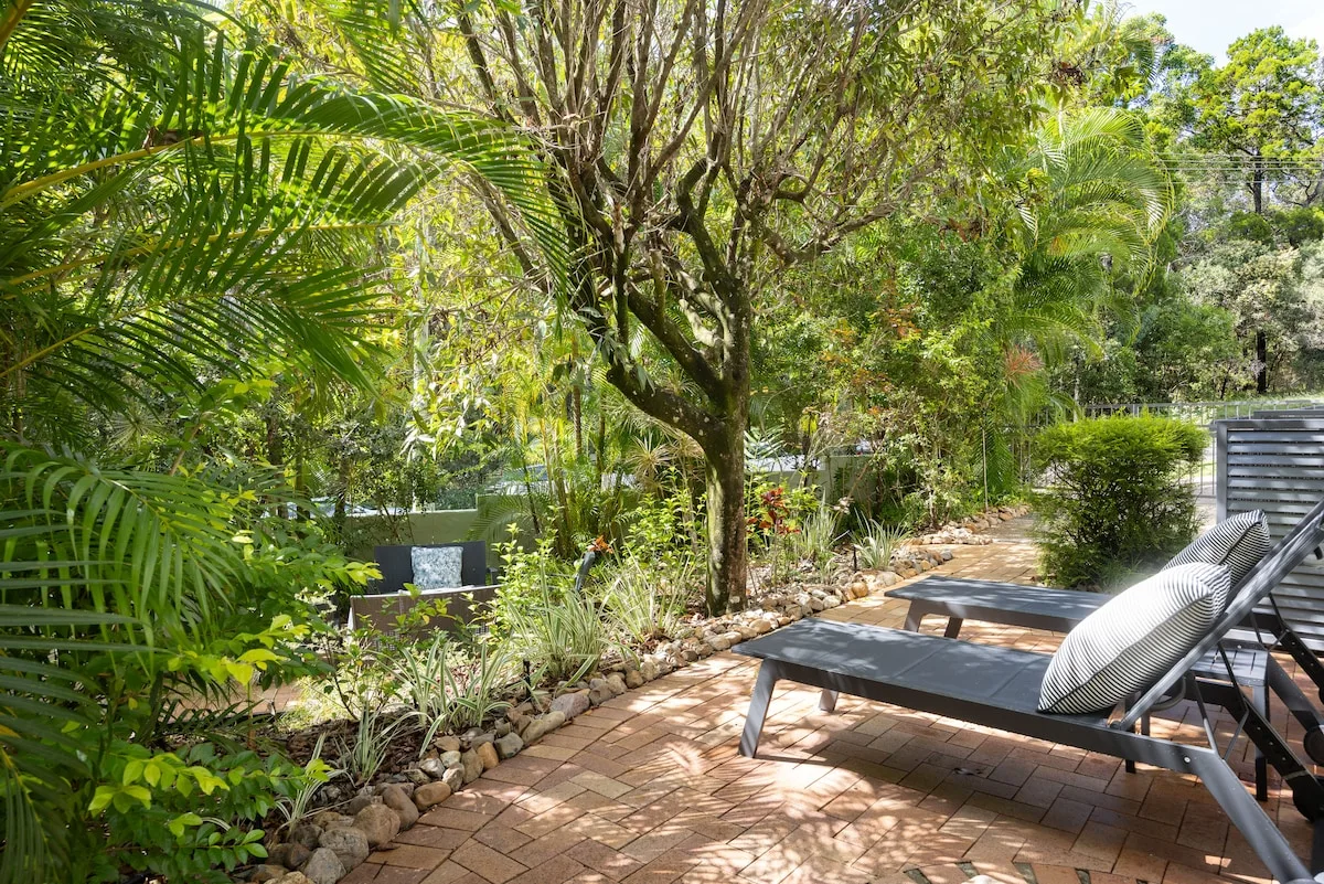 A leafy garden patio with lounge seating beneath trees and palms.