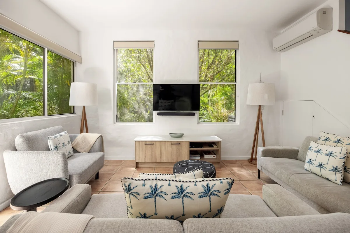 A bright living room with seating arranged around a television and large windows.