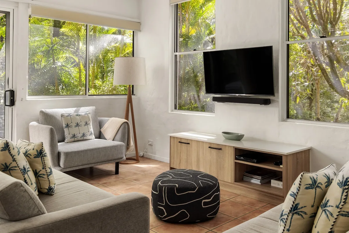 A bright living room with seating, a television, and large windows overlooking greenery.