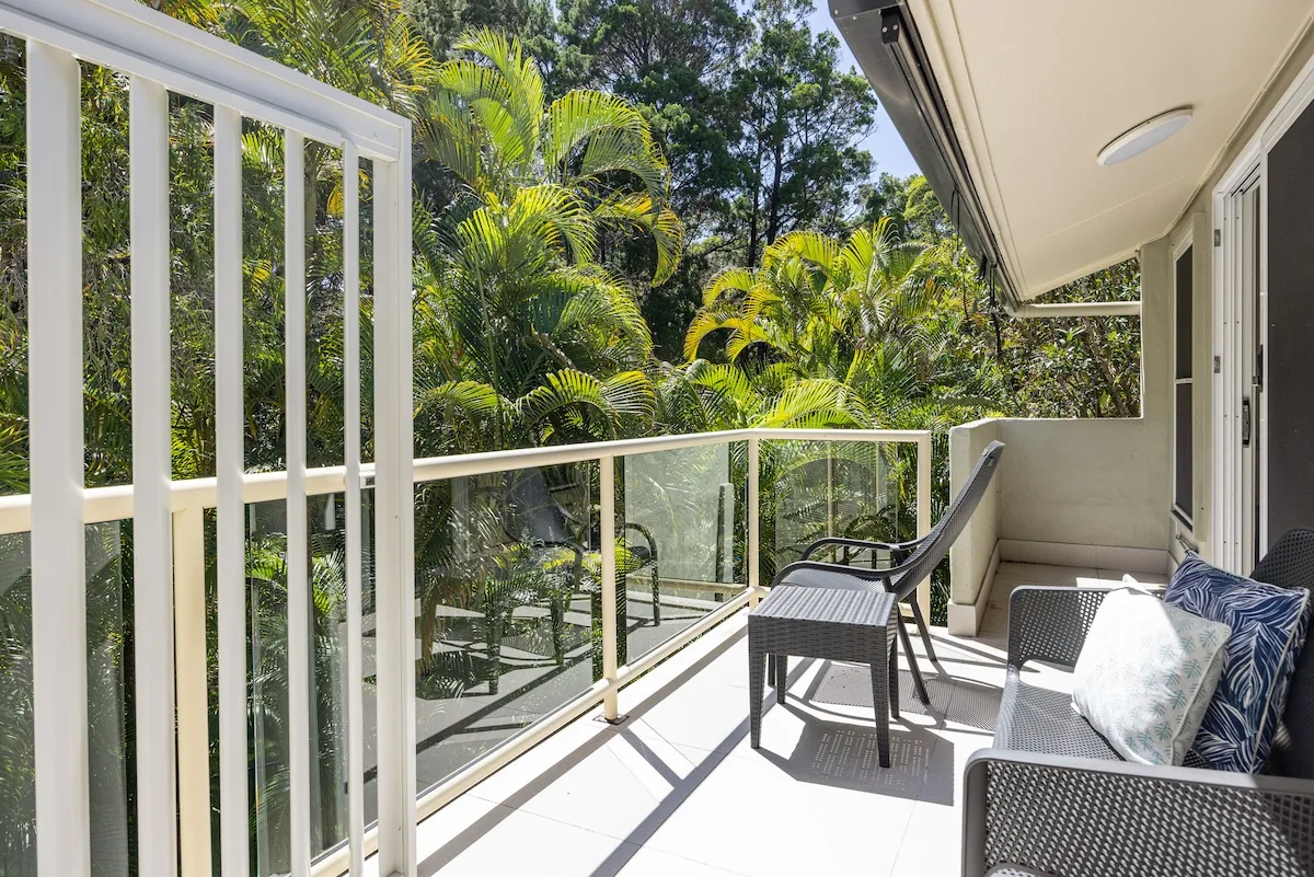 A balcony with outdoor seating overlooks lush green trees.