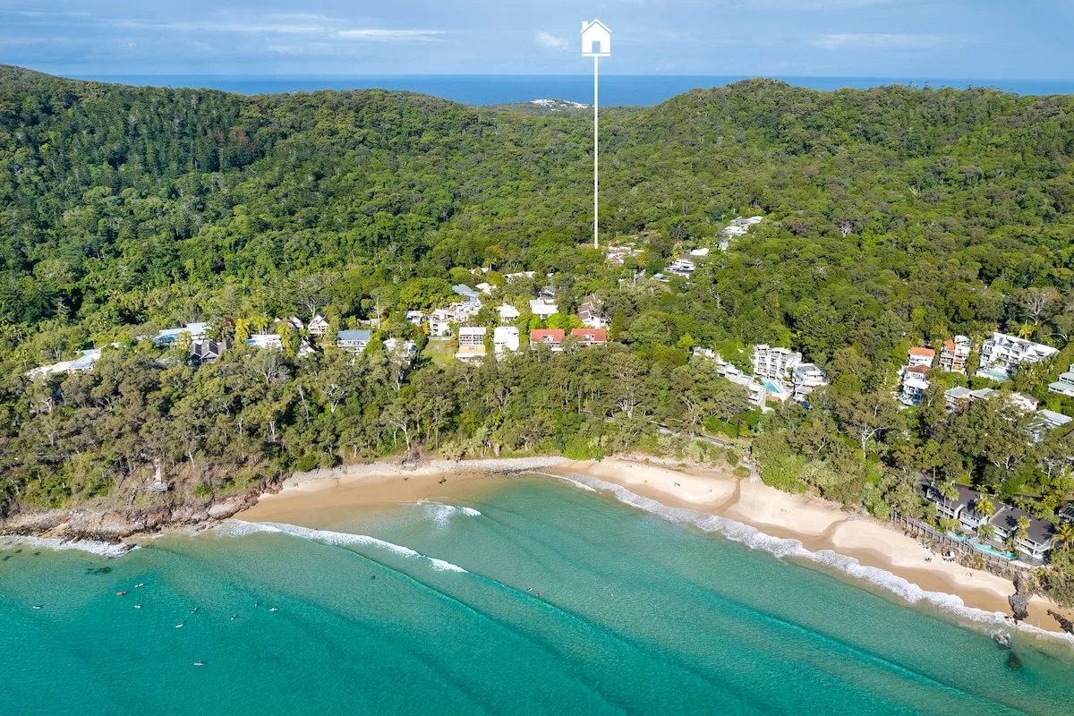 An aerial view shows a turquoise bay, sandy beach, and forested hills.