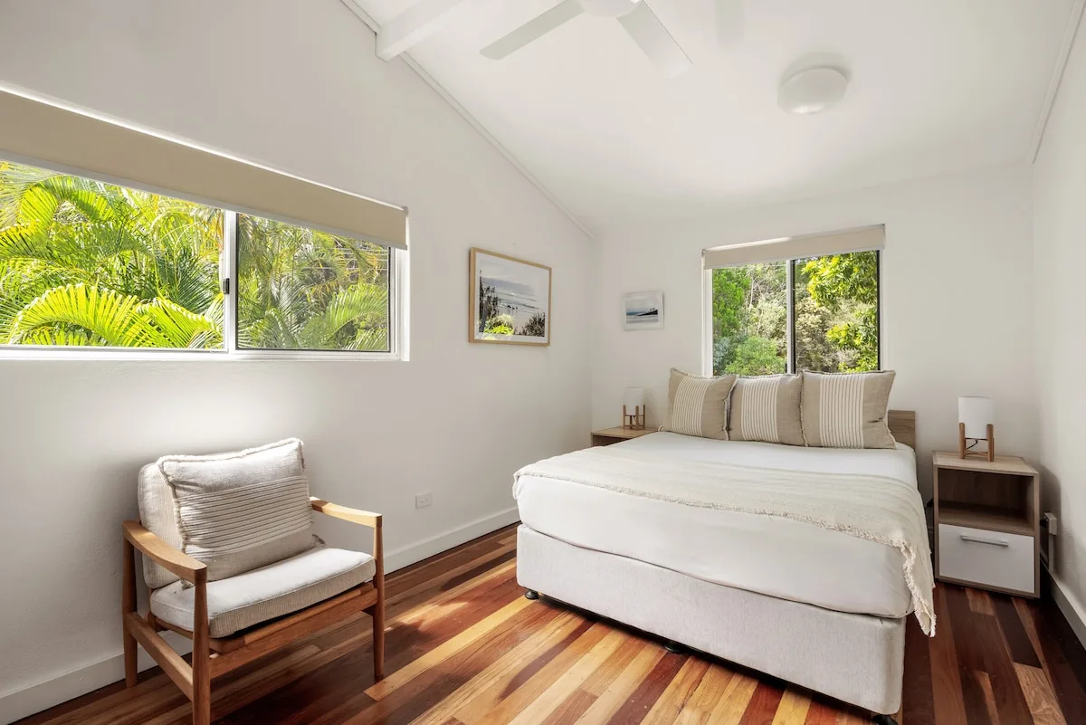 A bright bedroom with a bed, a chair, and windows overlooking greenery.