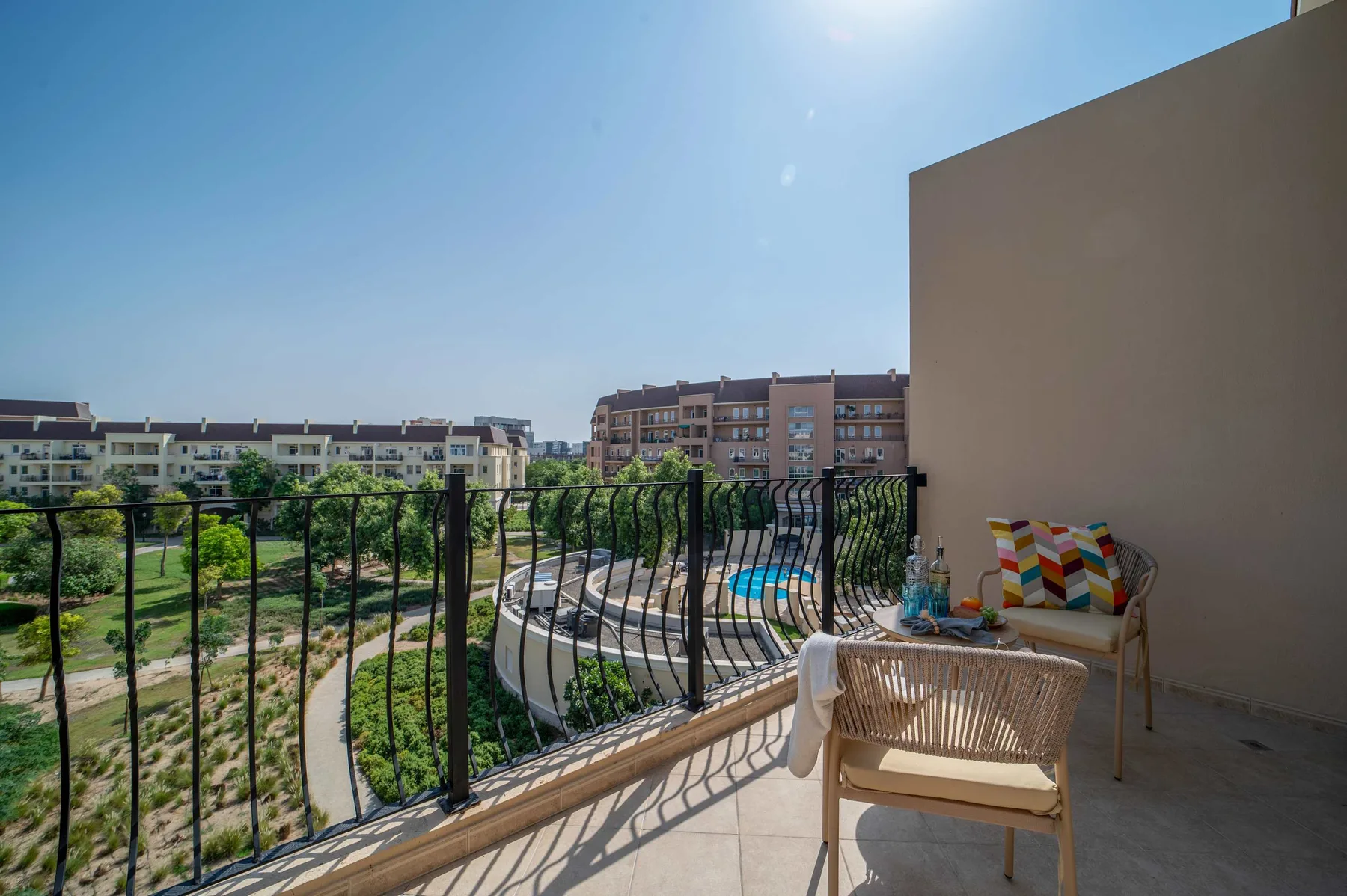 A sunny balcony with outdoor seating overlooks a landscaped courtyard and nearby buildings.