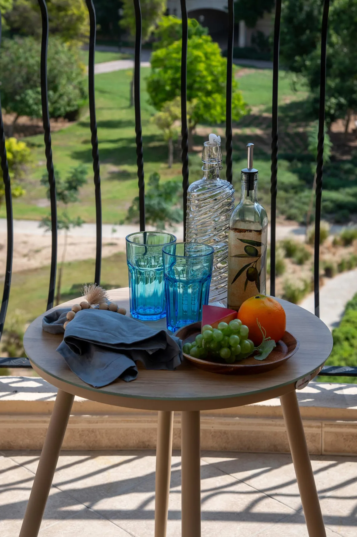 A small outdoor table holds blue glasses, fruit, and bottles beside a garden.