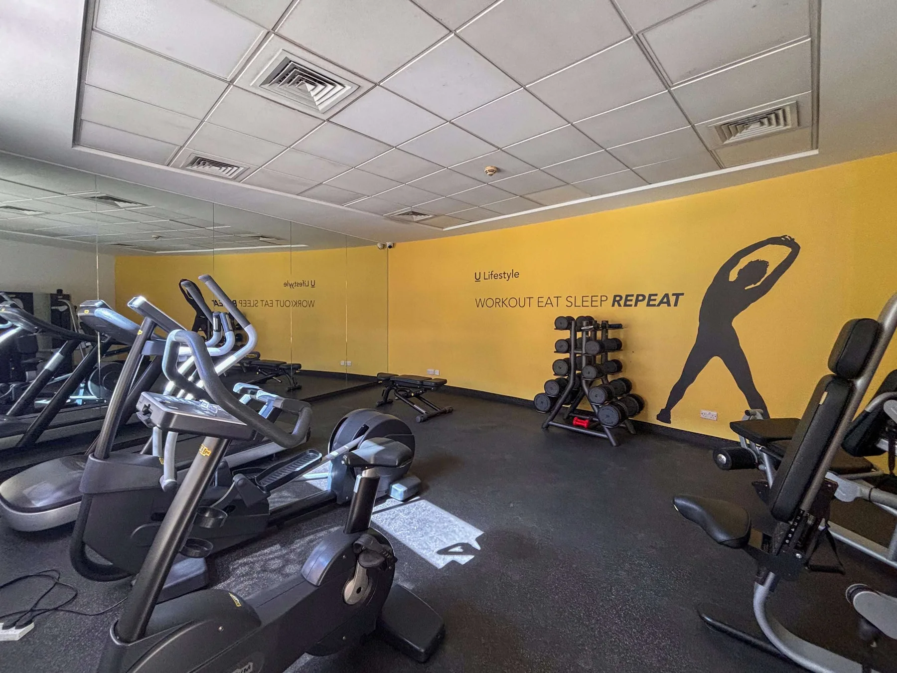 A fitness room with exercise machines and weights along a yellow wall.