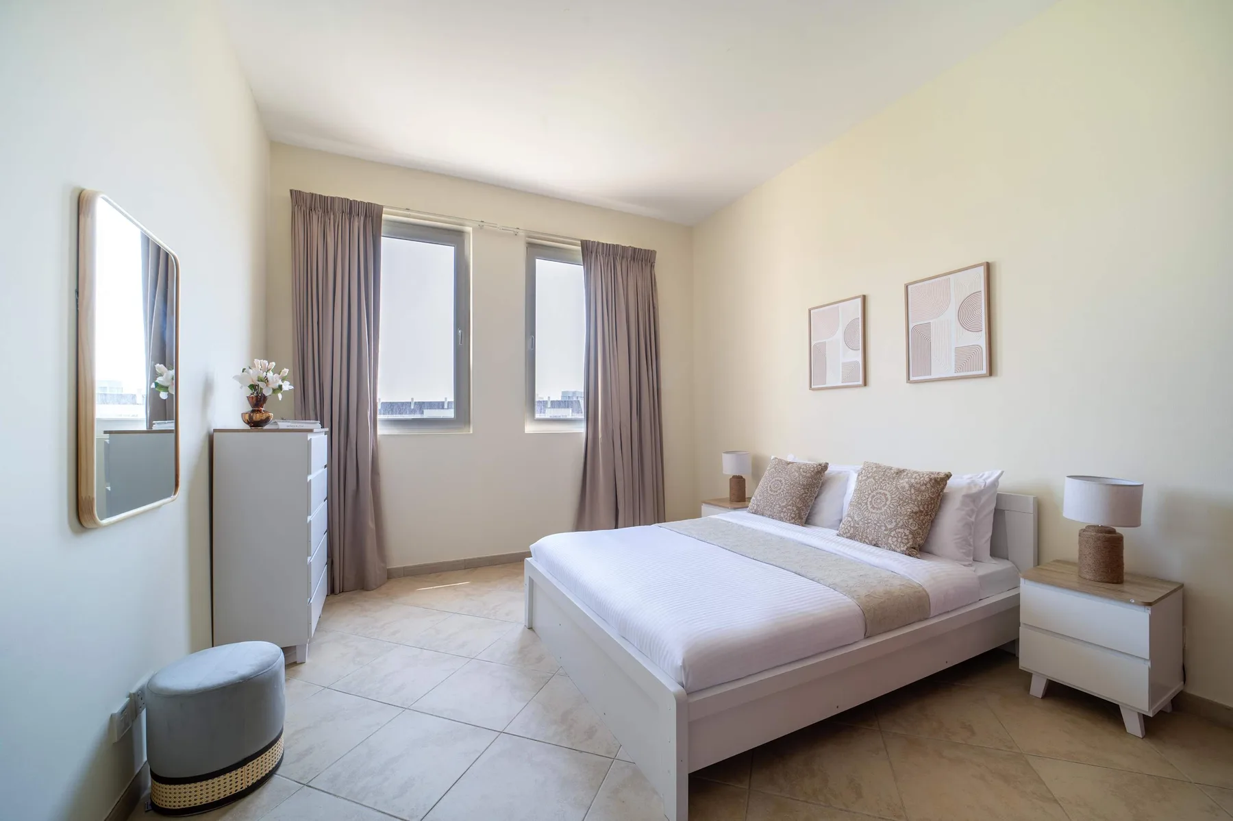 A bright bedroom with a neatly made bed, bedside tables, and tall windows.
