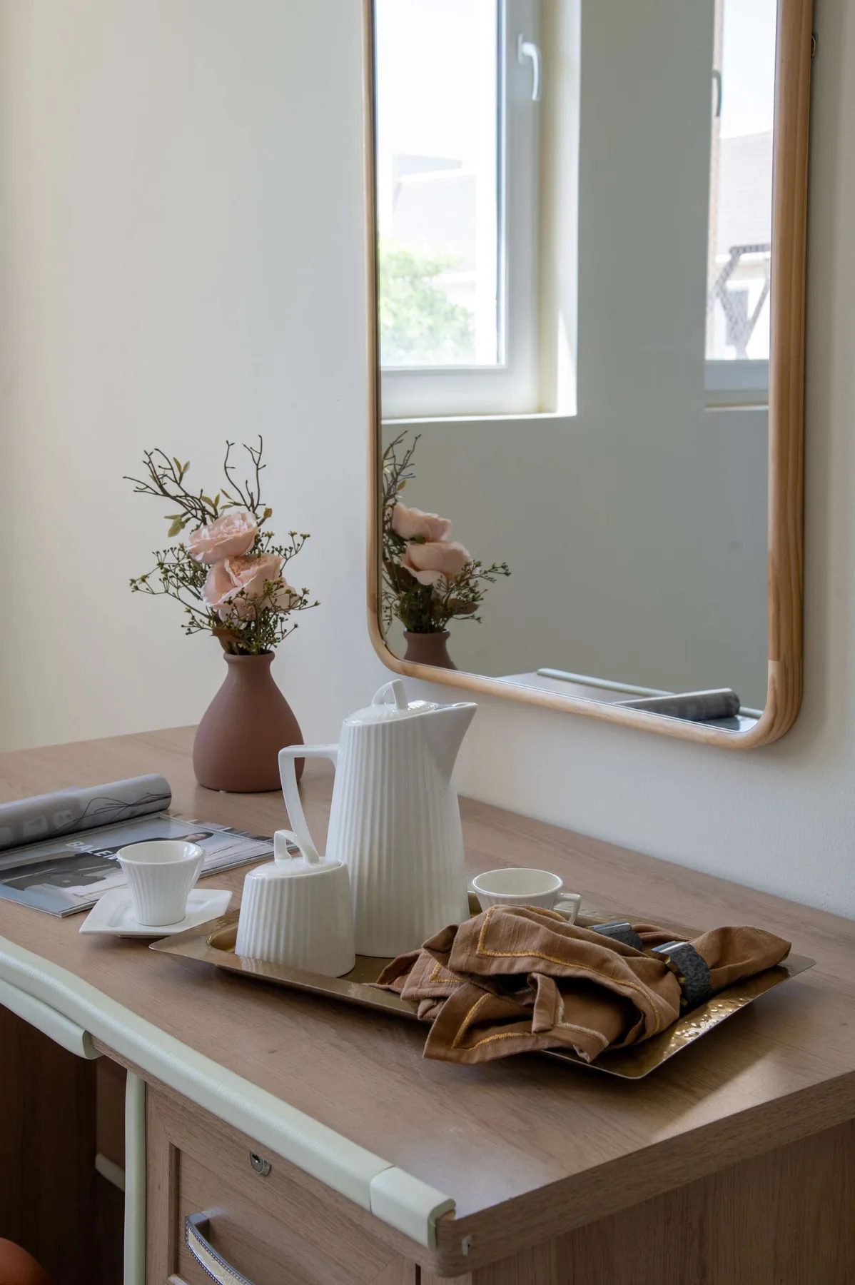 A wooden desk displays a white pitcher, cup, folded fabric, and a vase of flowers.