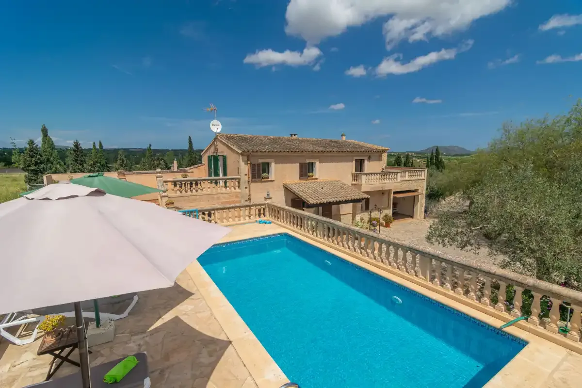 A stone terrace surrounds a bright blue swimming pool beside a countryside house.