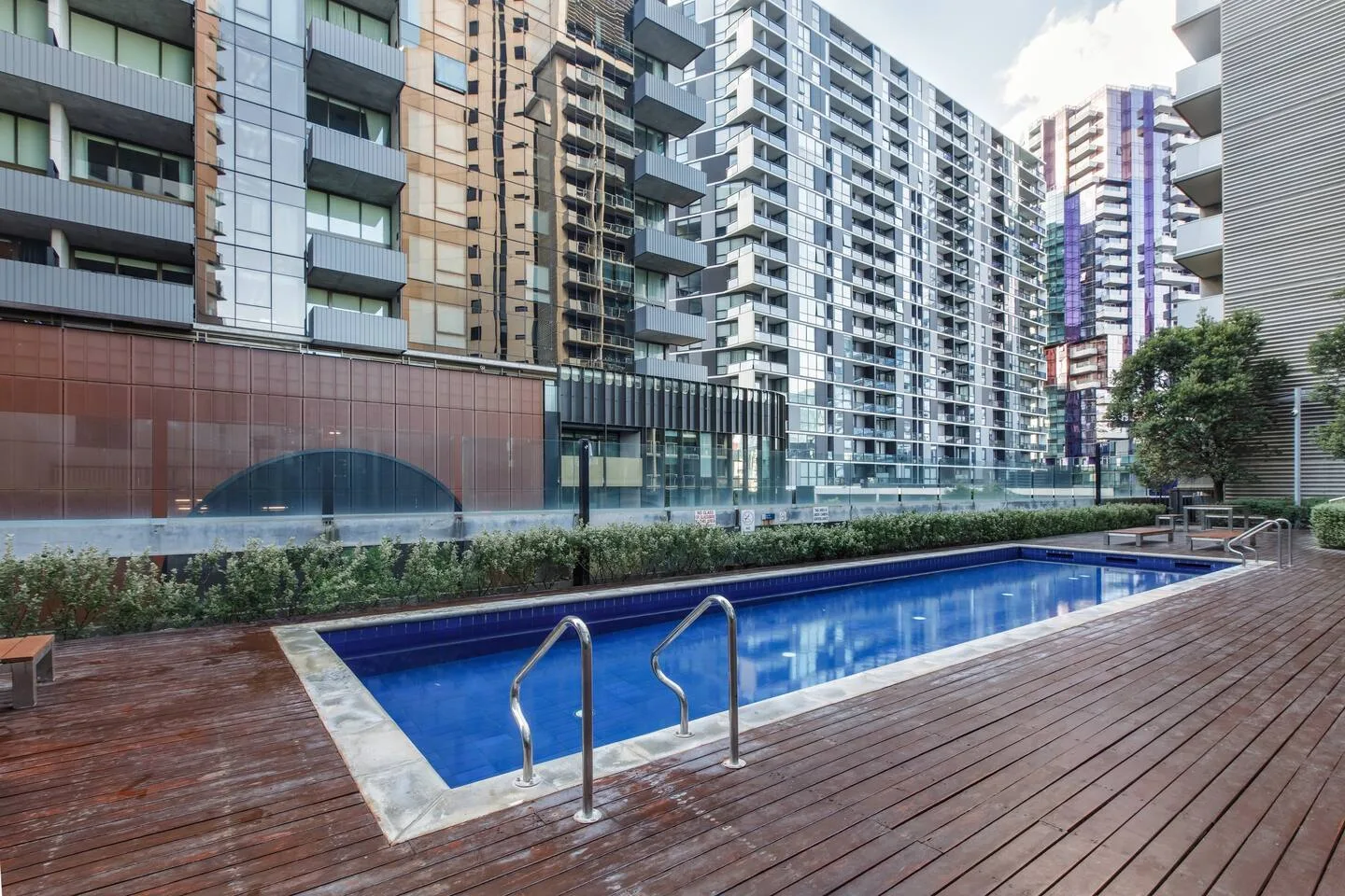 Una piscina al aire libre está rodeada de edificios altos, con una terraza de madera alrededor.