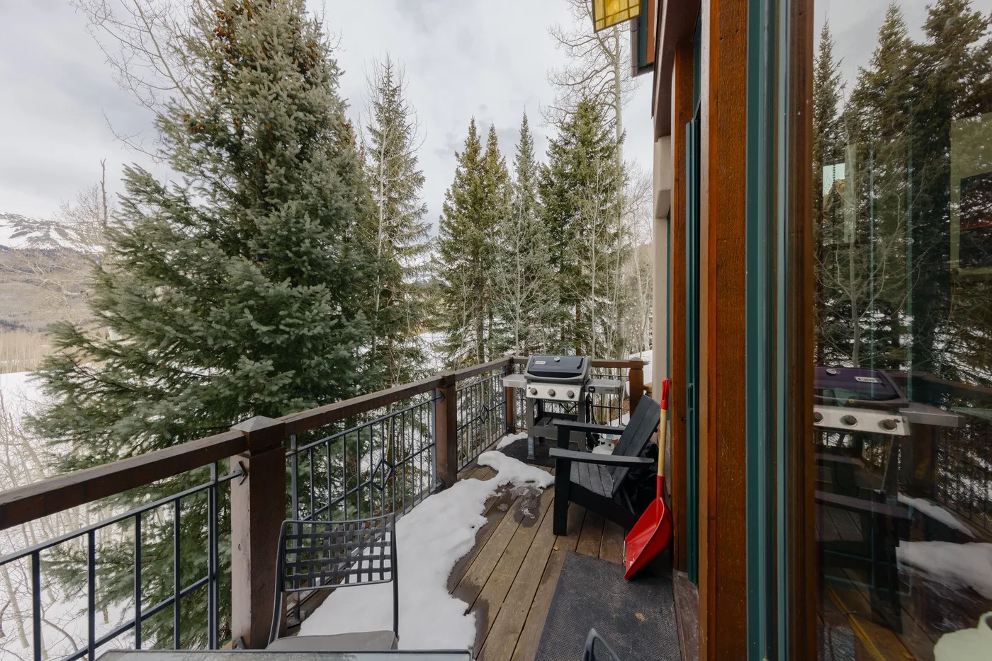 Una terraza de madera da a árboles nevados, con asientos y una parrilla junto a puertas de vidrio.