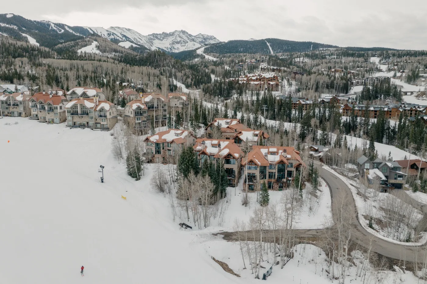Edificios de un complejo cubiertos de nieve junto a un camino sinuoso, con montañas al fondo.