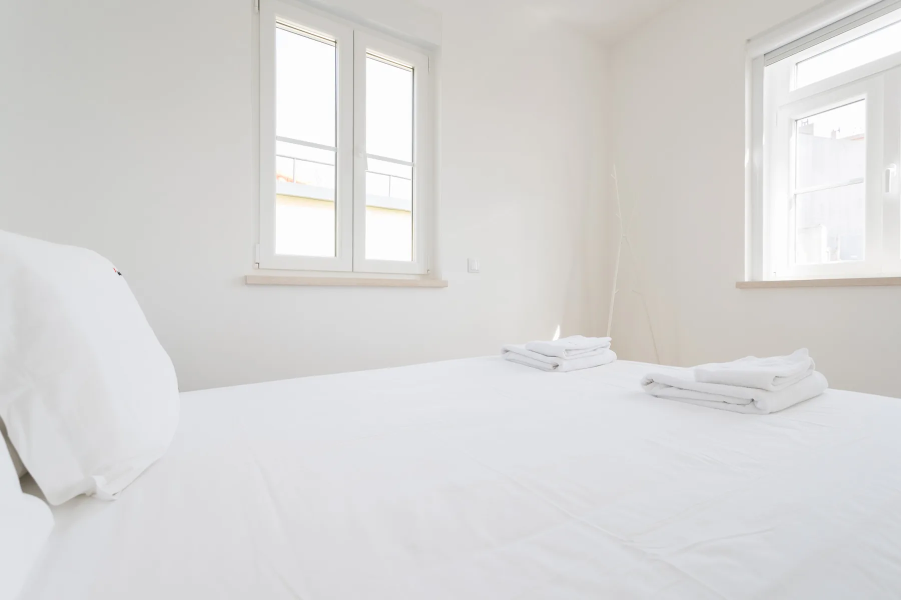 A bright bedroom with white bedding and two windows.