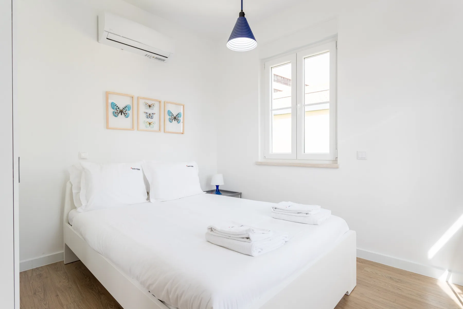 A bright bedroom with a white bed, framed butterfly prints, and a window.