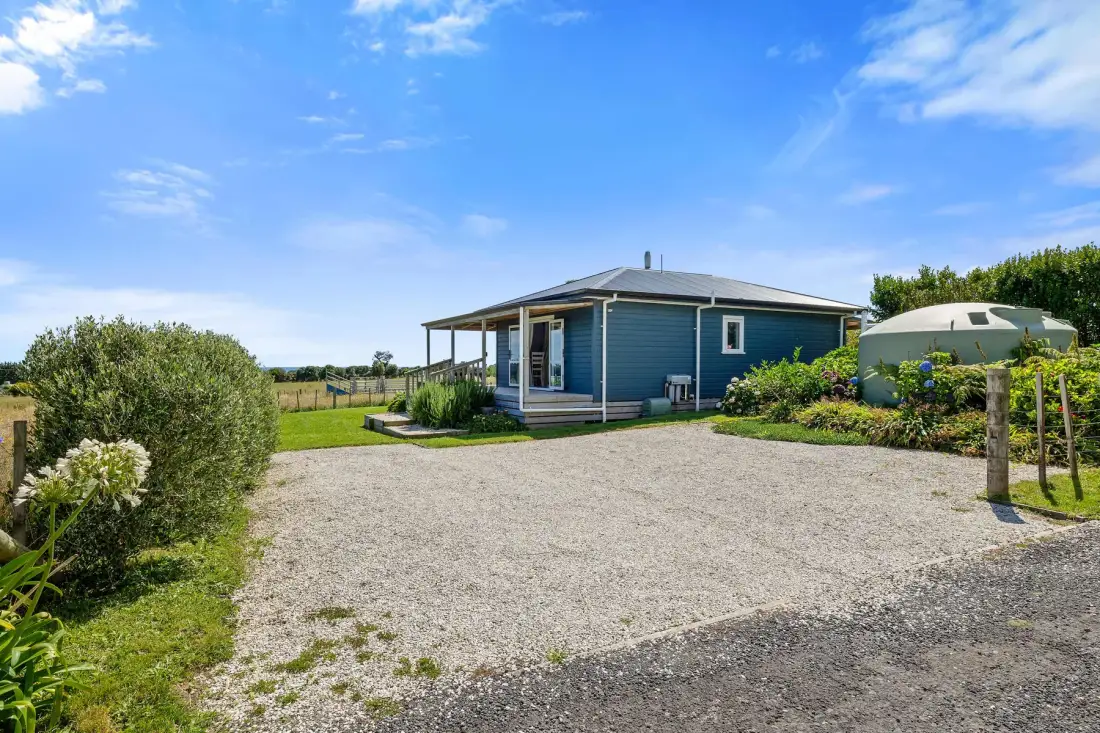 A blue cottage stands beside a gravel driveway and green lawn.
