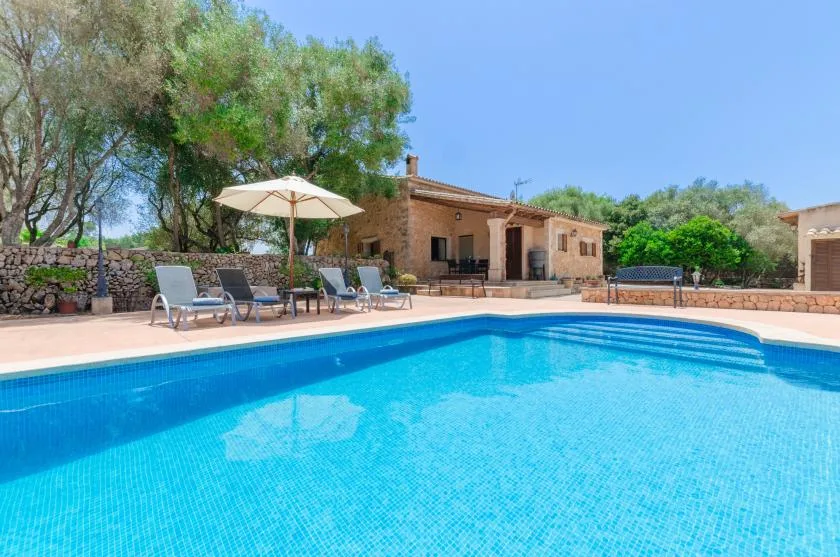 An outdoor swimming pool beside a stone house, with lounge seating under an umbrella.