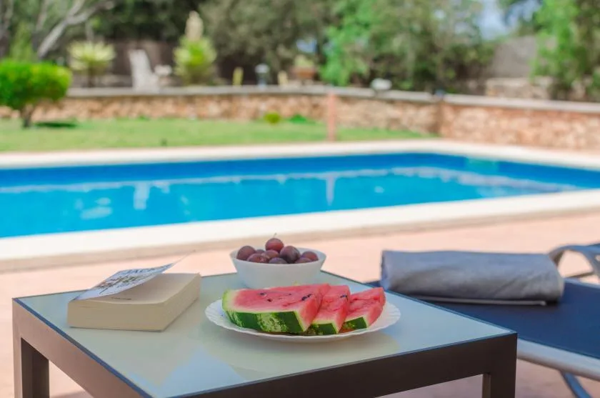 A poolside table holds a plate of watermelon and a bowl of fruit beside blue seating.