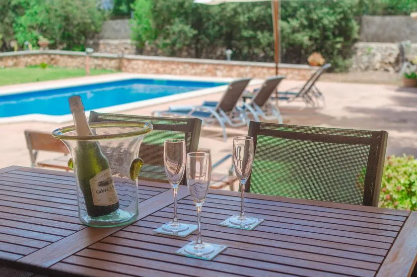 A poolside patio table is set with glasses beside a swimming pool.