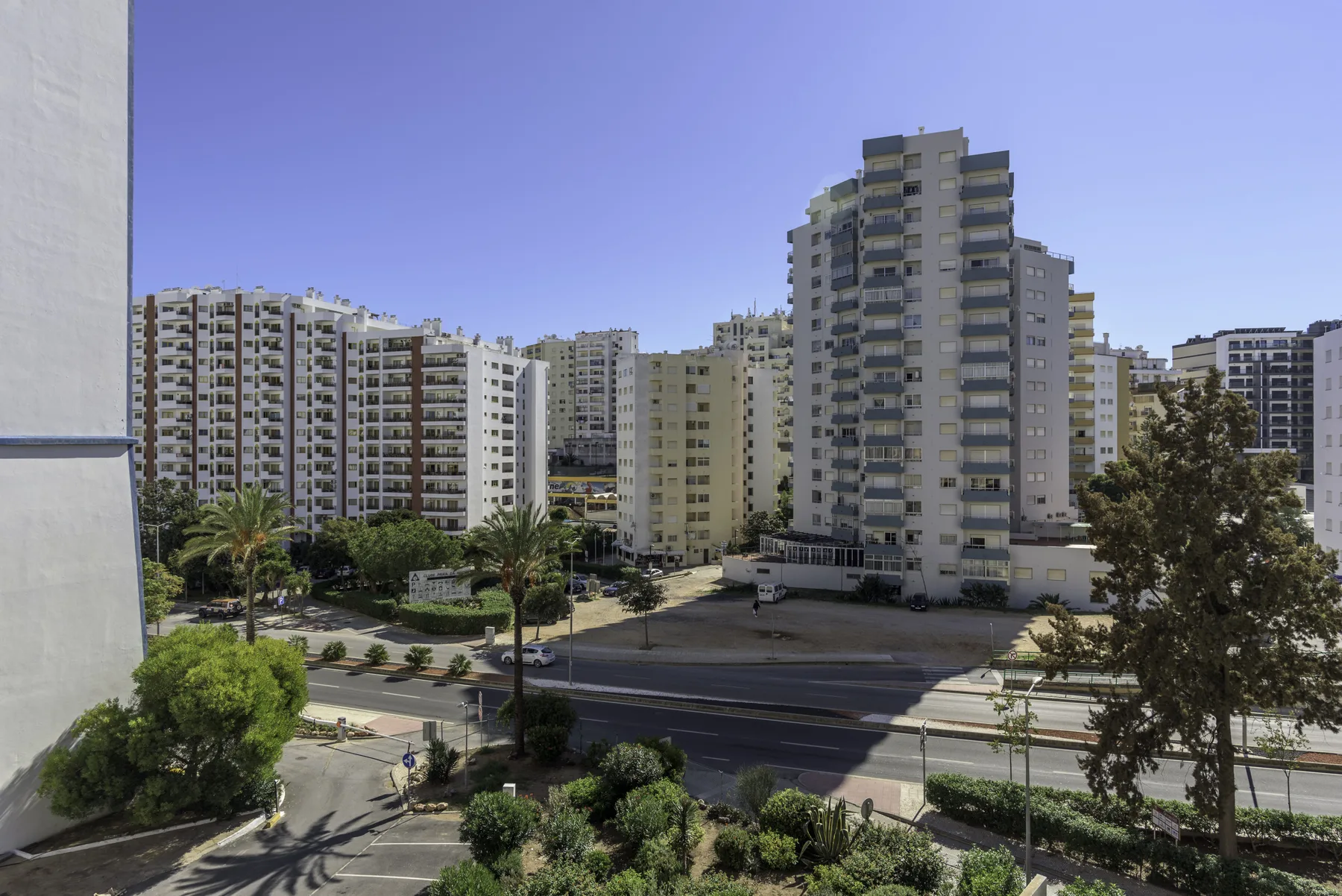 A city view from a balcony overlooks apartment buildings, a street, and greenery.