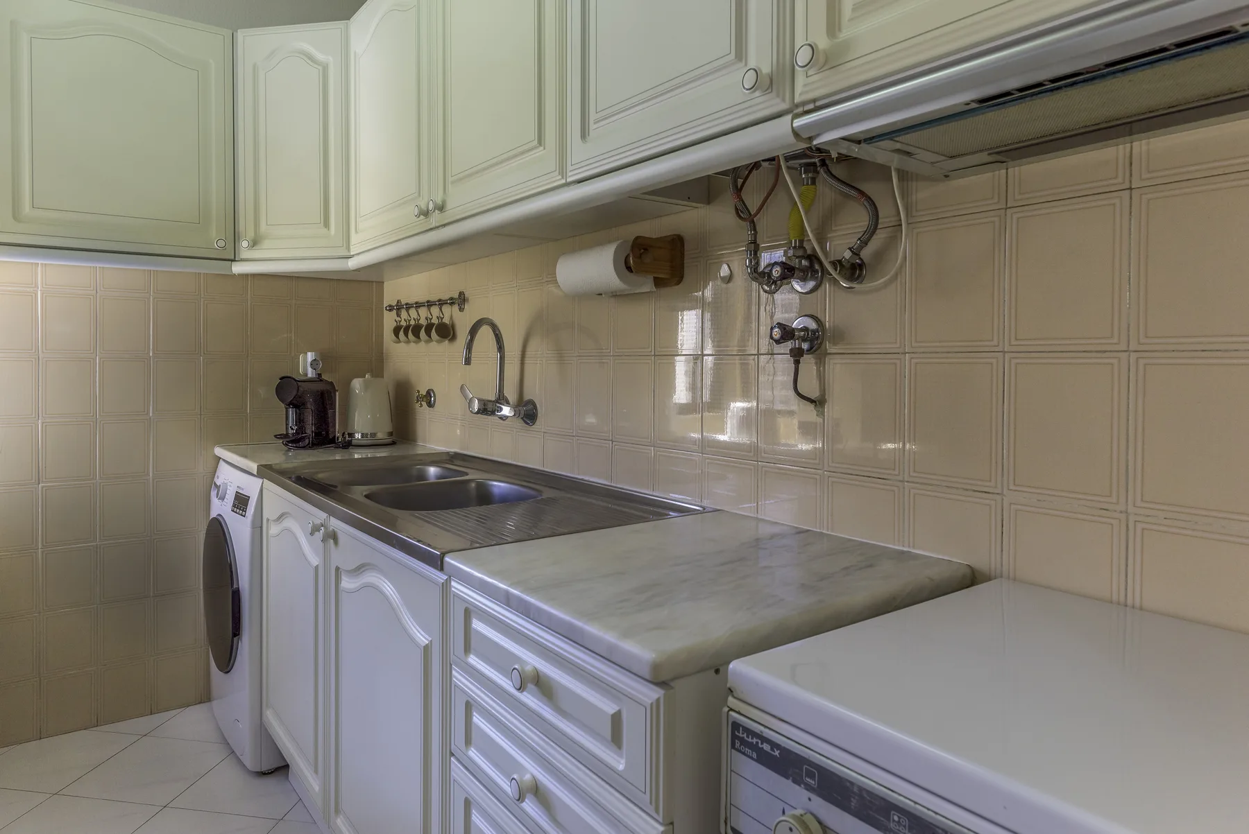 A tiled kitchen with pale cabinets, a sink, and laundry appliances.