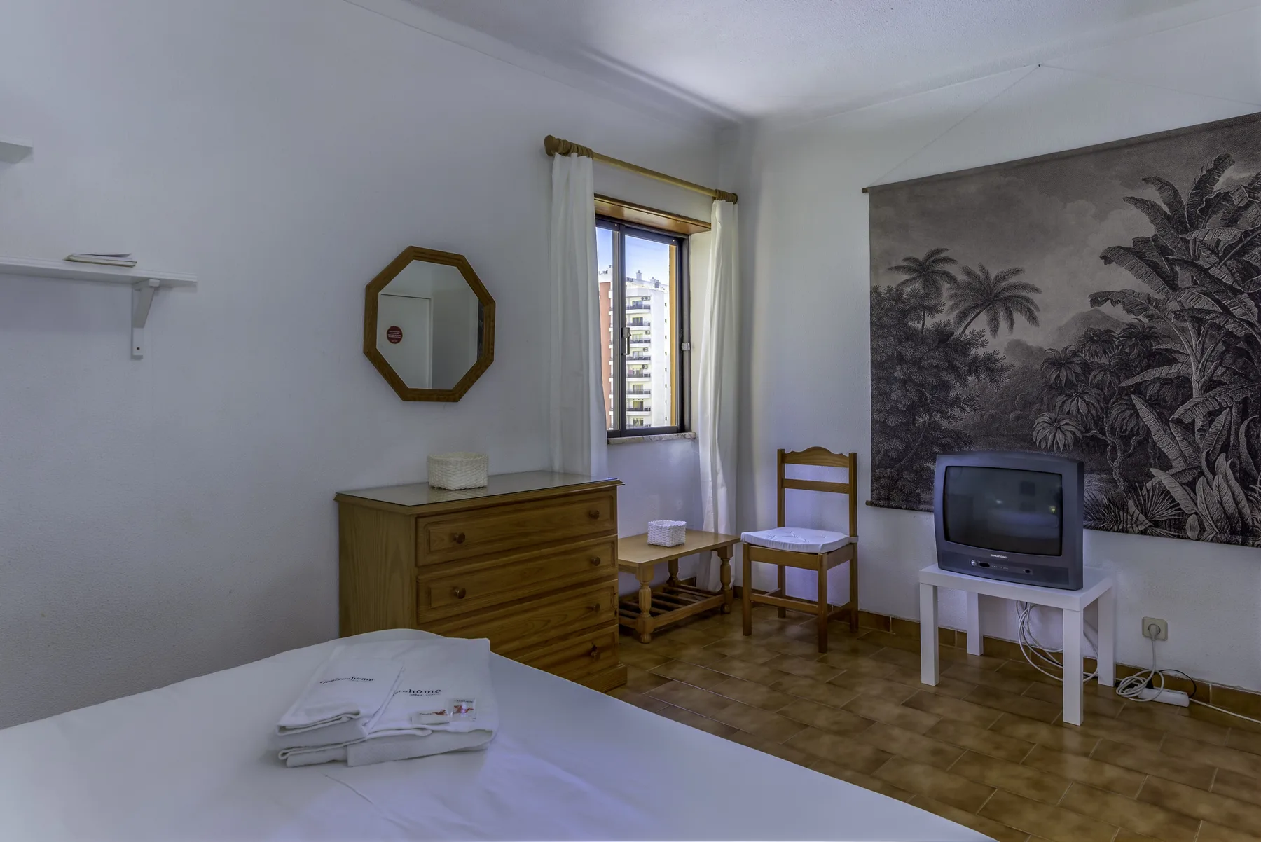 A bedroom with a dresser, a bed, and a television beneath a palm-tree wall mural.