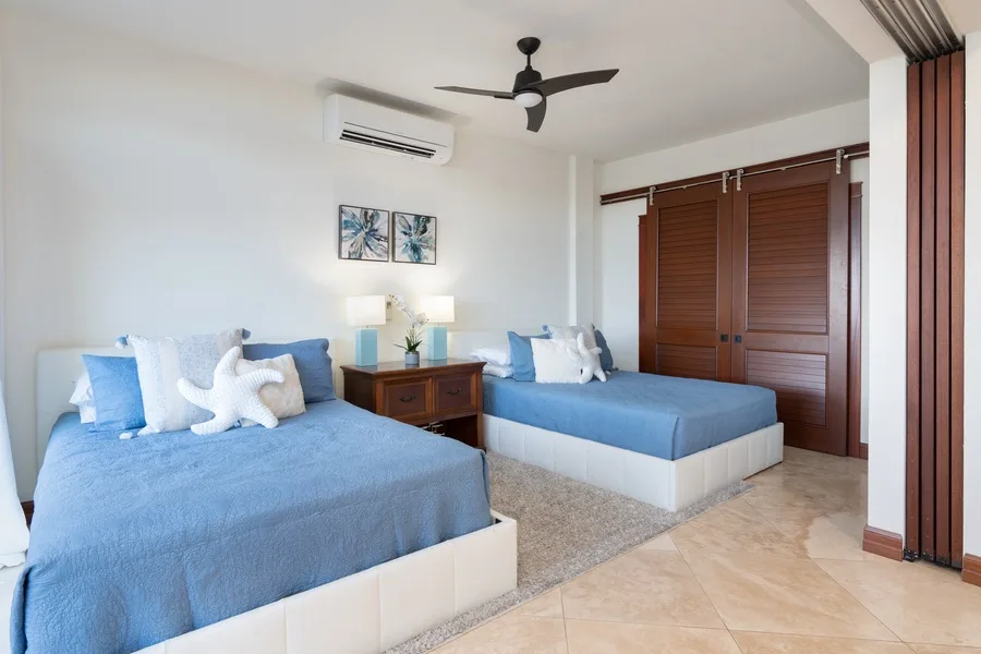 A bright bedroom with two beds, blue bedding, and wooden closets.