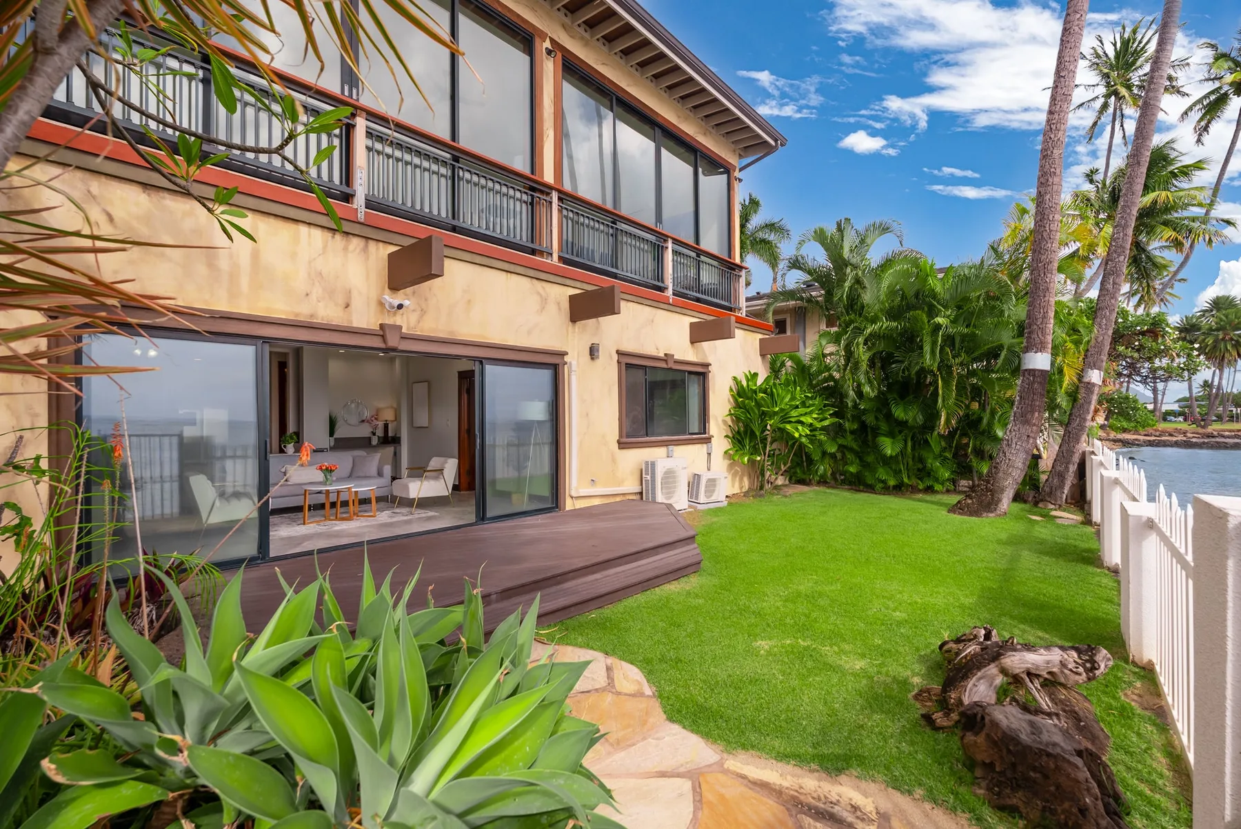 A two-story house opens onto a green lawn, with a patio and tropical plants.