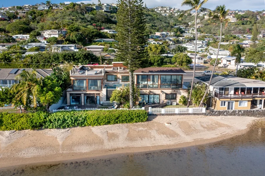 Coastal homes line a sandy shoreline, with a green hillside neighborhood behind them.