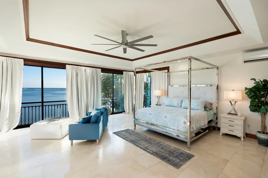 A bright bedroom with a canopy bed, blue seating, and ocean views through large windows.