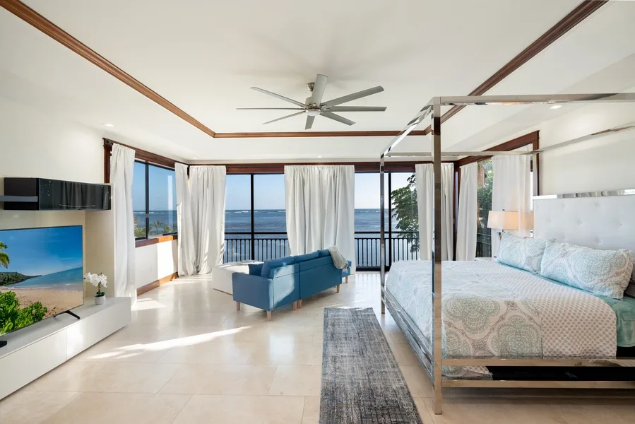 A bright bedroom with a bed, seating area, and ocean views through large windows.