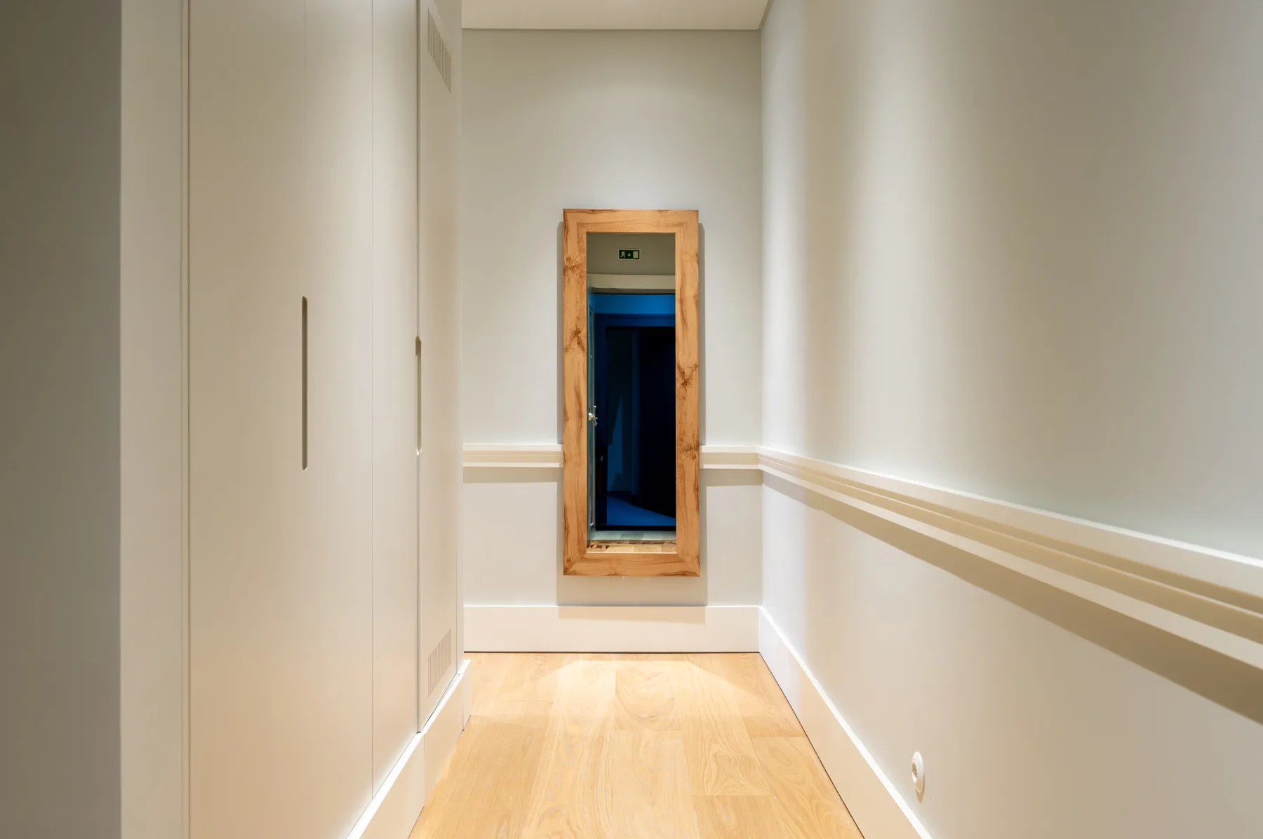 A narrow hallway with light wood flooring and a mirror at the end.