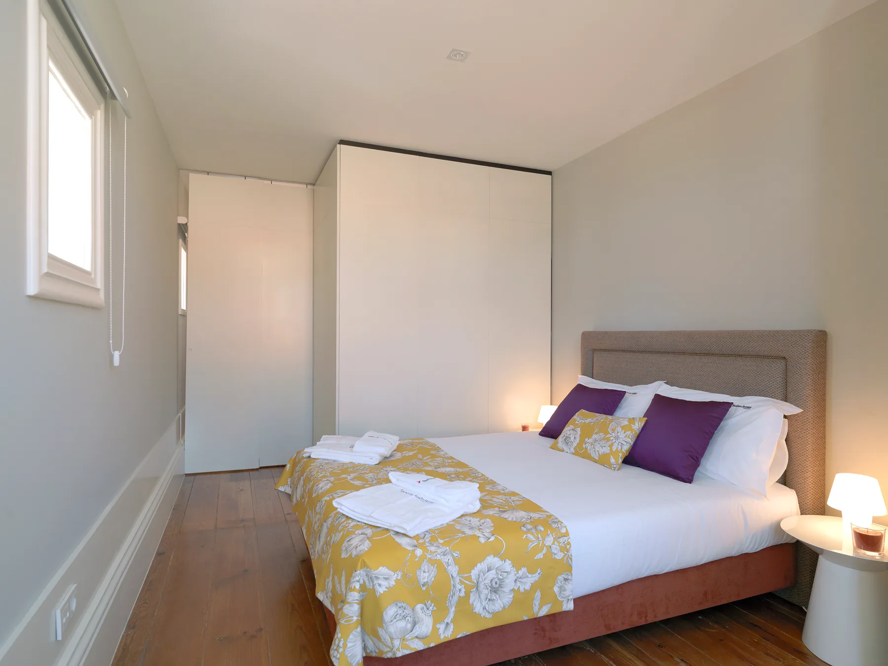 A bedroom with a bed dressed in white linens and a yellow patterned cover.