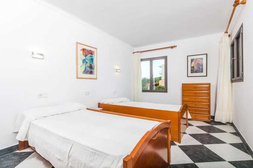 A bright bedroom with two beds, a wooden dresser, and windows.