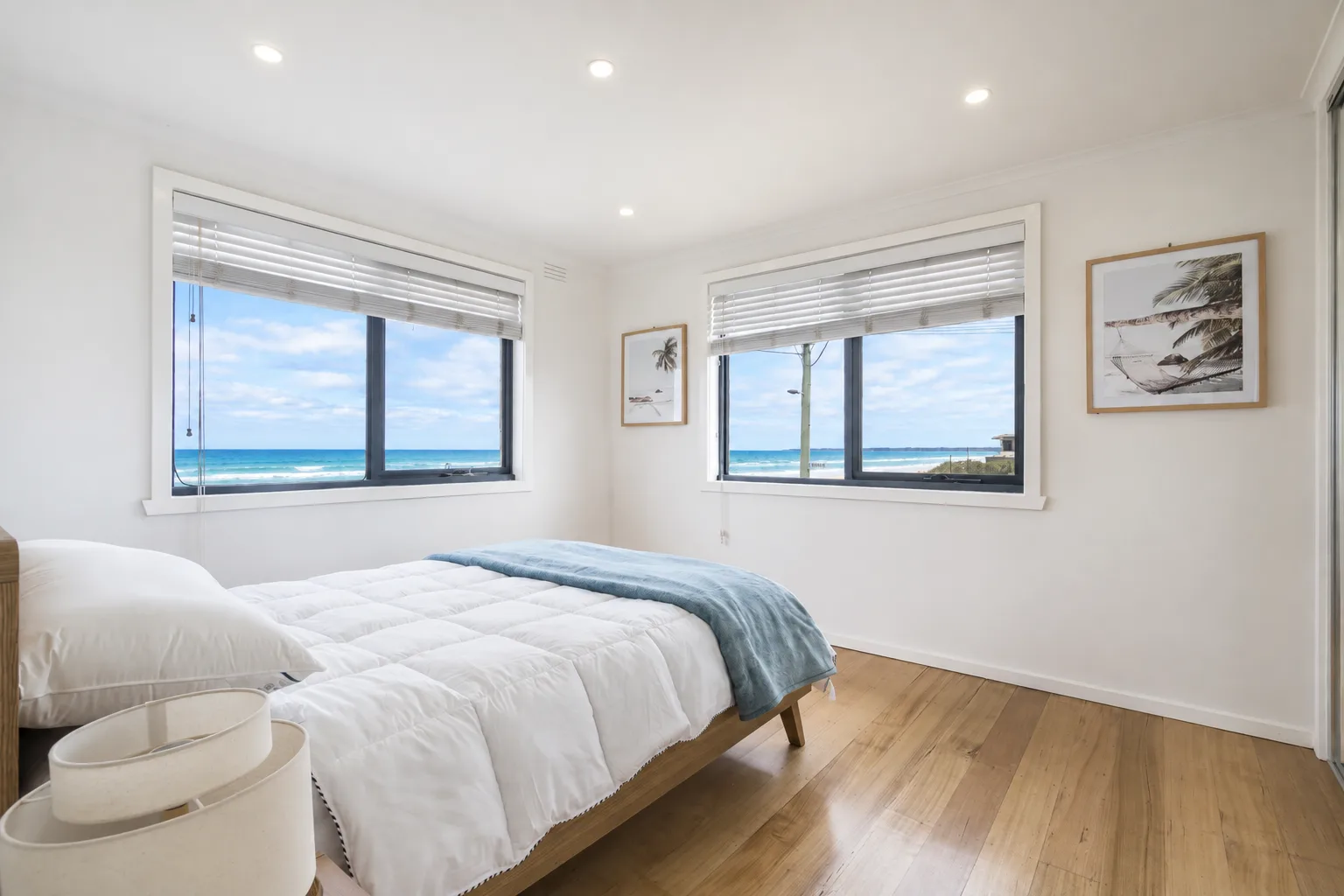 A bright bedroom with a bed beside two windows overlooking the sea.