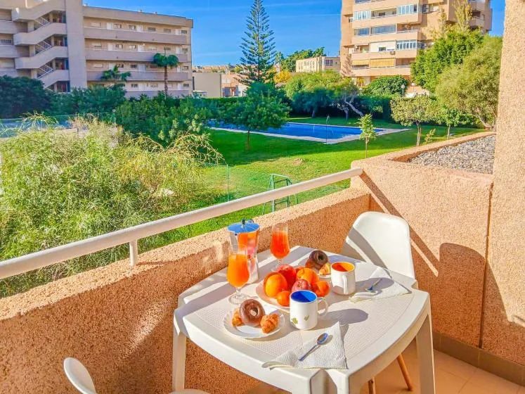 Un balcón soleado con una pequeña mesa de comedor da a un patio ajardinado y una piscina.