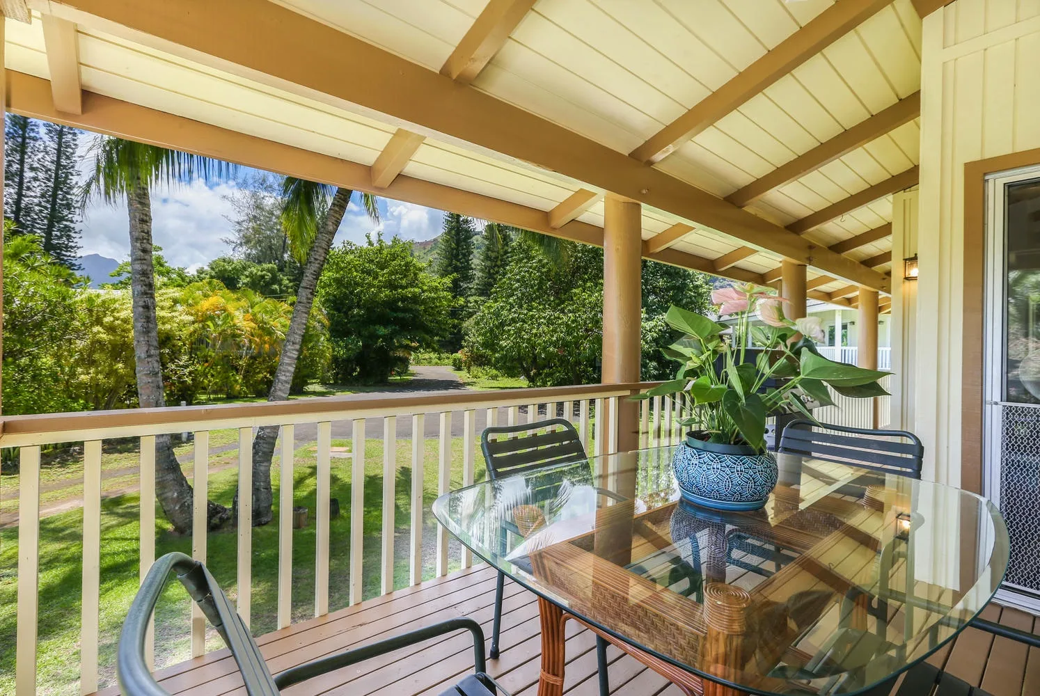 Un patio cubierto tiene una mesa de vidrio y asientos junto a un jardín frondoso.