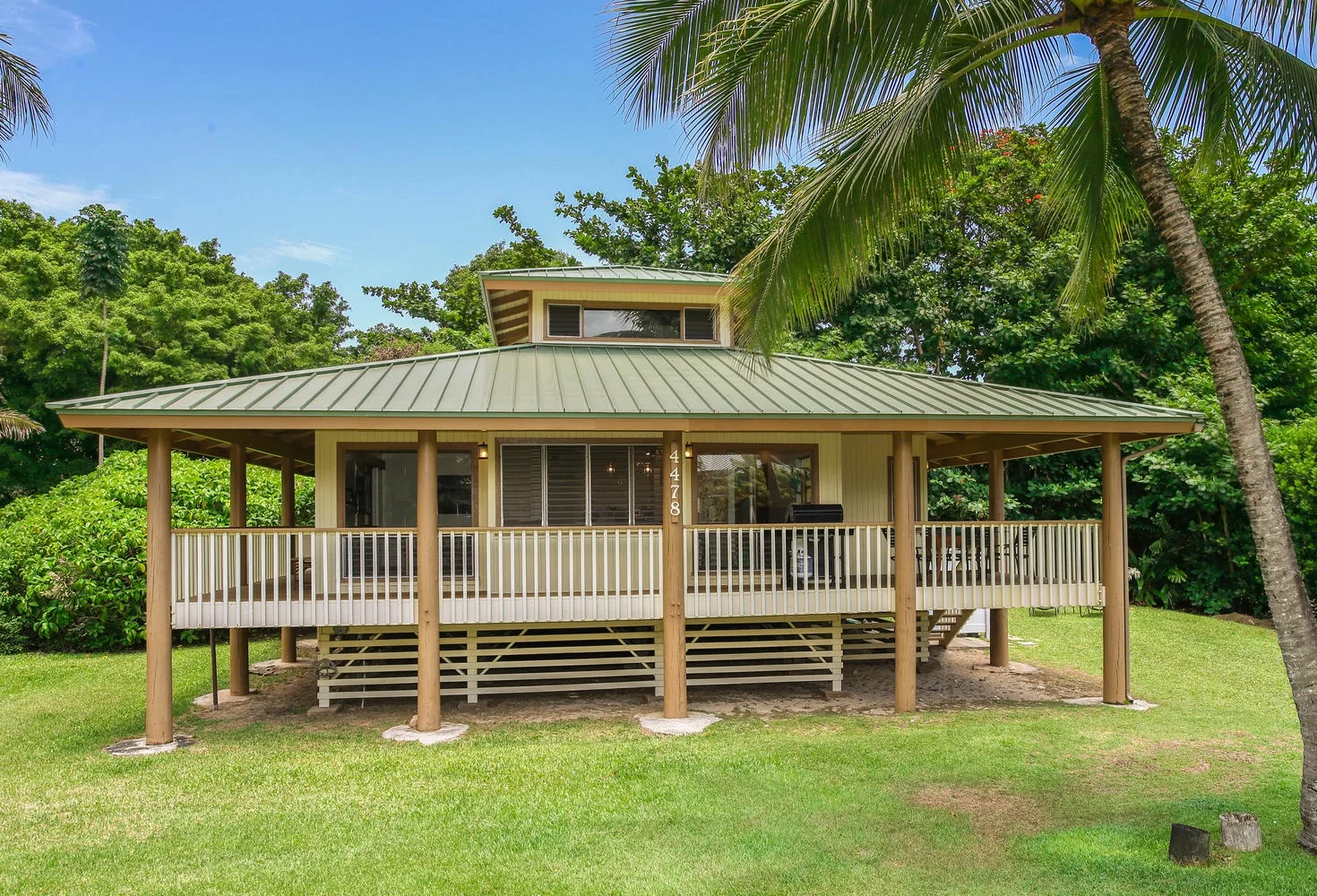 A raised house with a broad covered porch stands amid green trees and lawn.