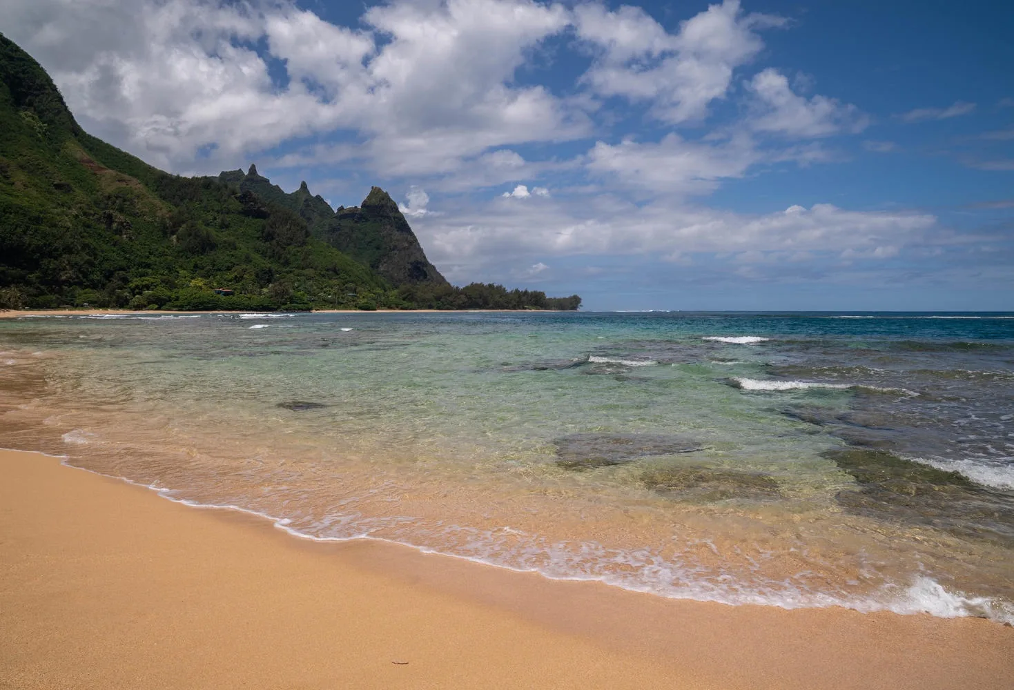 A sandy beach meets shallow turquoise water beneath a green, rugged coastline.