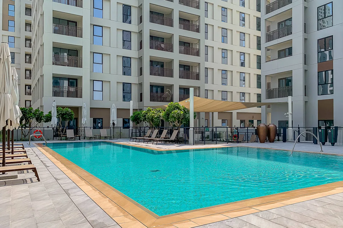 An outdoor swimming pool sits among tall apartment buildings, with lounge seating along the deck.