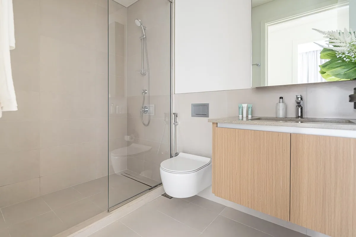 A bright bathroom with a glass shower, wall-mounted toilet, and wood-toned vanity.