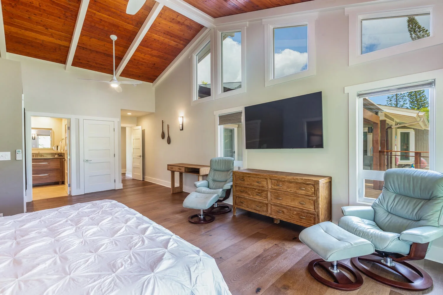 A bright bedroom with a bed, a television, and seating beneath a vaulted wood ceiling.
