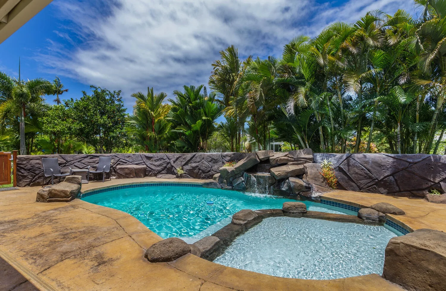 An outdoor swimming pool and spa sit among tropical palms and greenery.