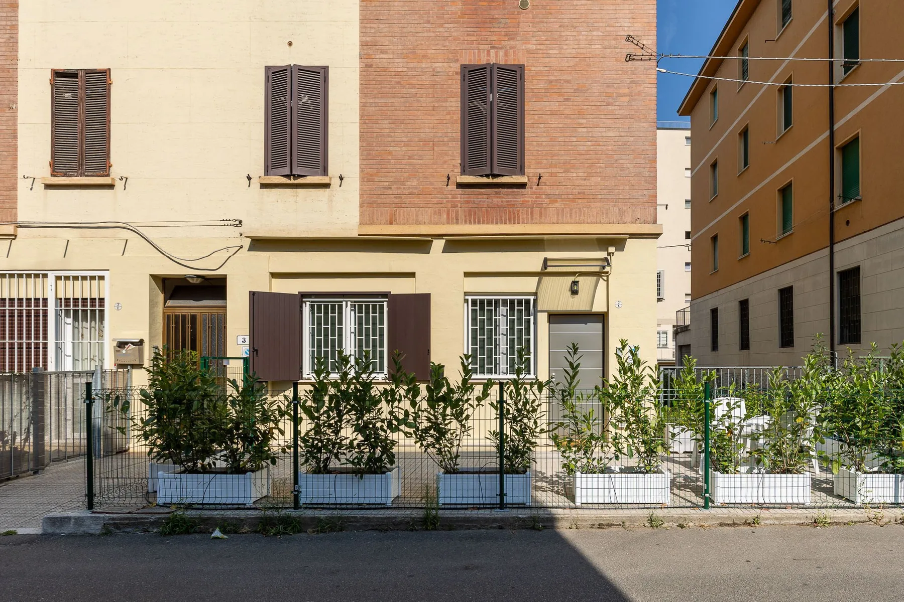 Street view of a residential building with shuttered windows and a fenced garden.